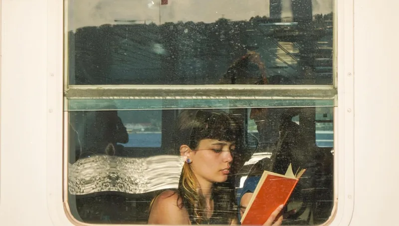 Solo female traveler with brown hair reading a red book by the window on a ferry during her trip.