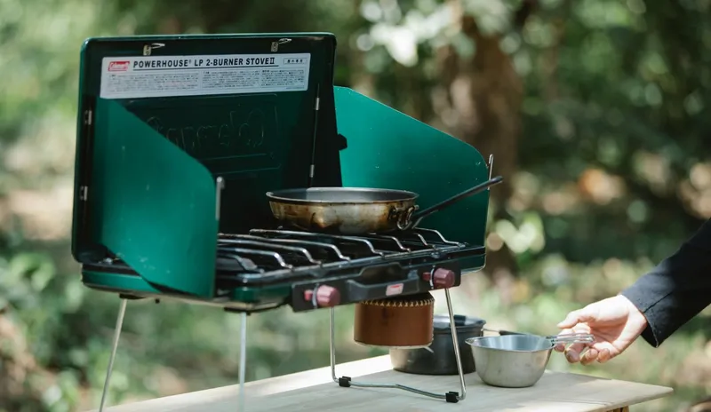 Outdoor cooking in nature with a green two-burner portable stove and a frying pan.