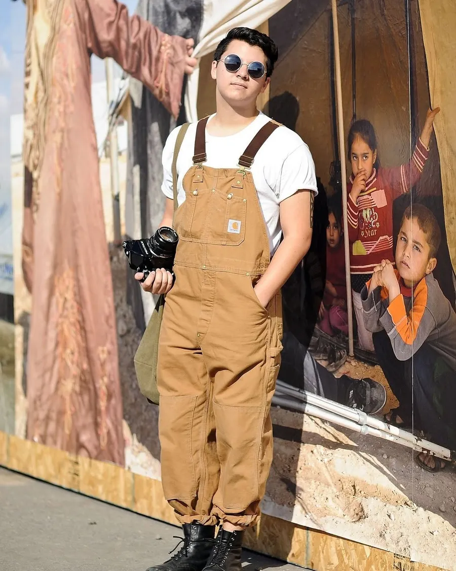 A man with sunglasses holding a camera and wearing brown Carhartt overalls and black boots/