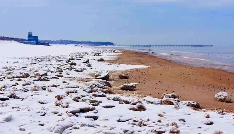 A snowy, rocky beach with a wavy shore, brown sand and a blue structure in the background.