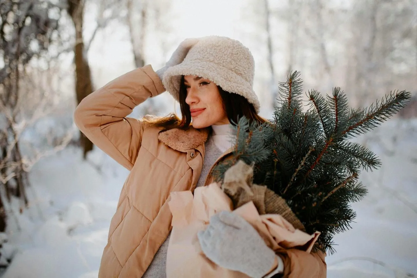 Young woman wearing a brown puffer jacket, a white hat and gloves carrying a foldable Christmas tree in the winter.