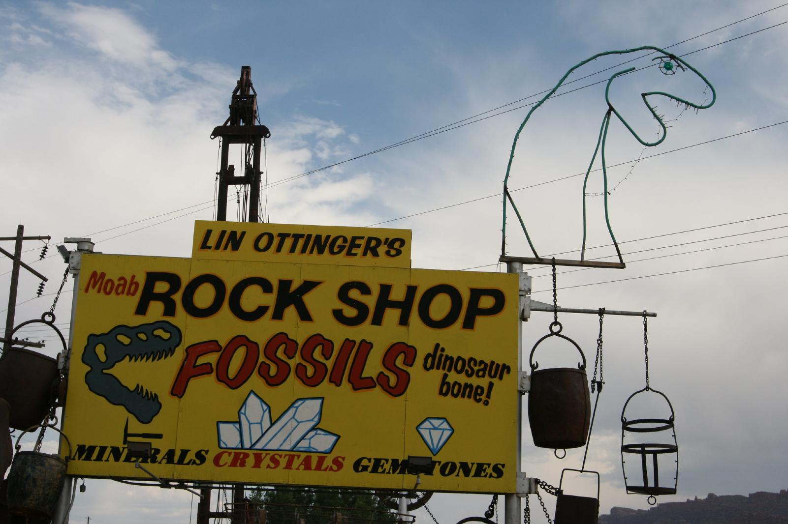 A yellow billboard for the Moab Rock Shop in Utah with dinosaur models and hanging buckets.