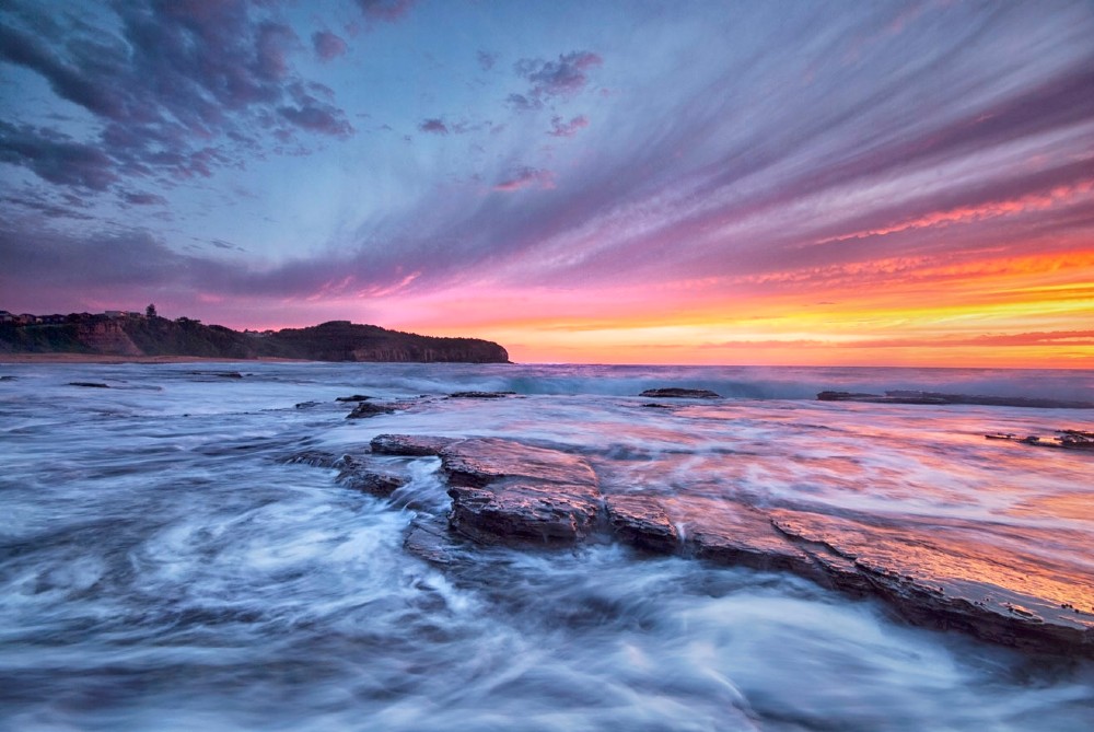 An orange and purple sky during a sunrise over a rocky shore at Narrabeen Beach in Australia.